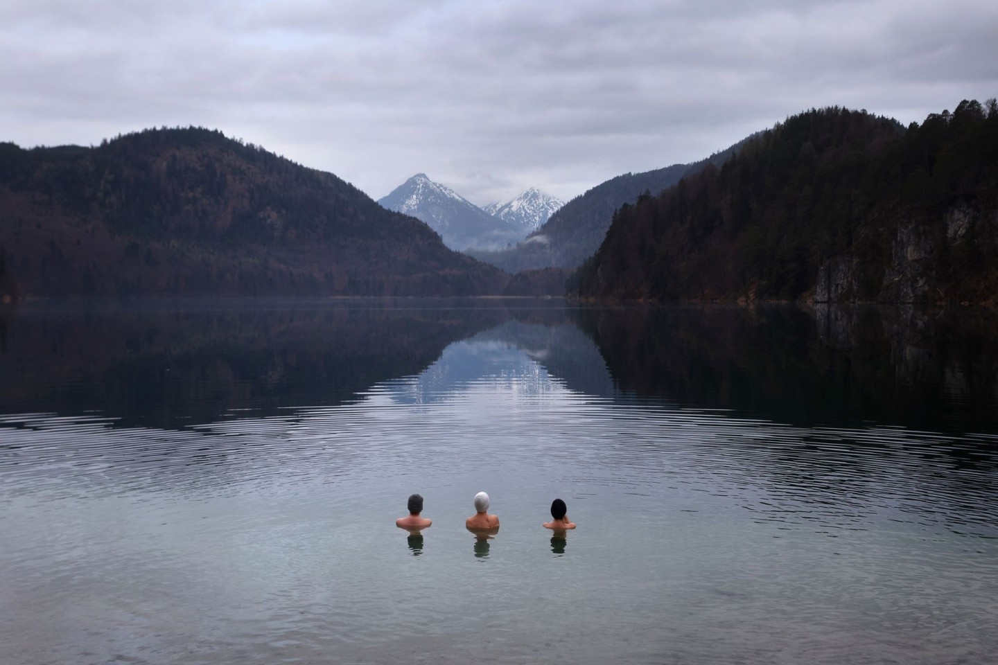 Drei Frauen baden am frühen Morgen unter wolkenverhangenem Himmel im noch kalten Wasser des Alpsees.