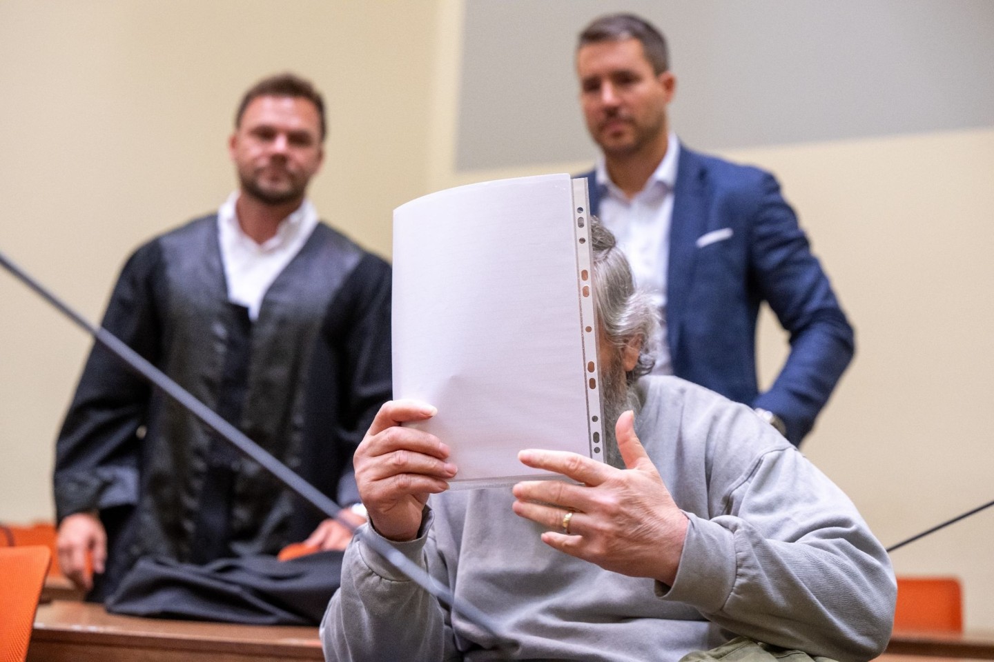 Der Angeklagte hält sich im Gerichtssaal ein Blatt Papier vor das Gesicht. Im Hintergrund stehen seine Anwälte.